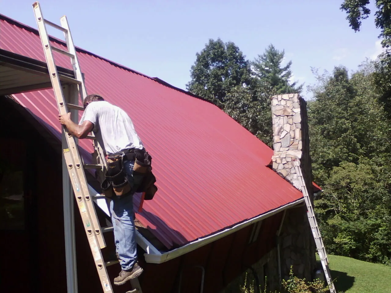 Roofer on a ladder beside a red metal roof and stone chimney — Soffit & Fascia Repair by Roofing Brothers in Schneider